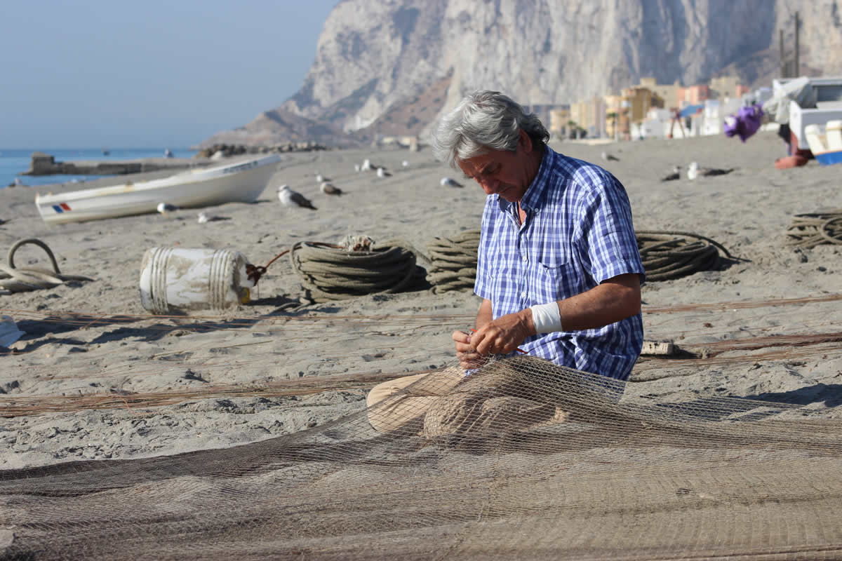 José  remienda el arte
Después de haber terminado el lance y haber  repartido la venta entre los marineros, el patrón, José, aprovecha la mañana  para remendar el arte. Las betas, el bidón, los cabos… se esparcen por la  playa, ocupan su espacio, troquelando un paisaje que es difícilmente compatible  con los usos derivados del turismo. José y su tripulación pueden ser  considerados los últimos mohicanos de un modo de vida en la playa, que se  inicia de madrugada y termina antes del almuerzo. Saberes heredados que siguen  sirviendo para garantizar una economía de subsistencia a un cada menor grupo de  gente de la playa del mediterráneo andaluz. (Autora: Gema Carrera, IAPH).